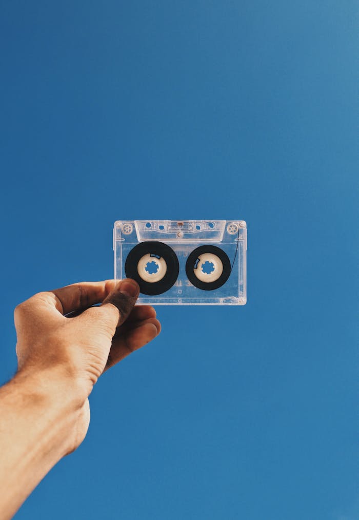 A hand holding a transparent vintage cassette tape against a clear blue sky, capturing nostalgia.